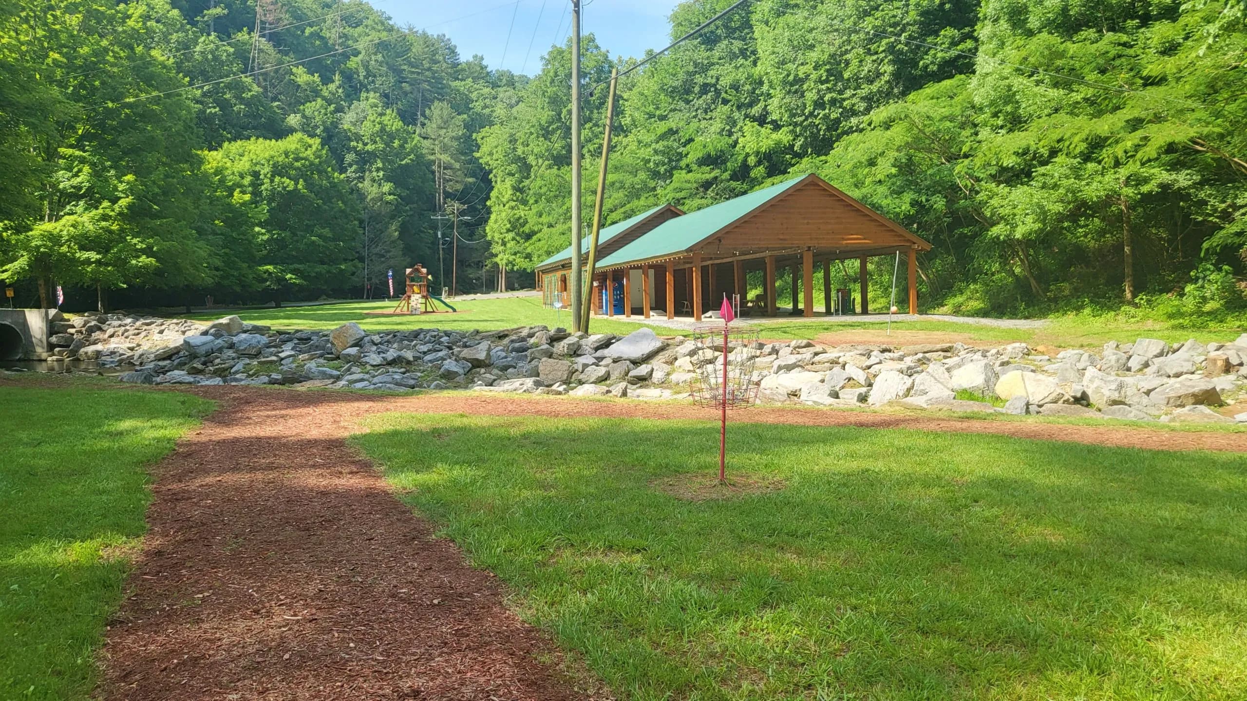 A disc golf basket on a lawn near a wooden open-air pavilion and a rock-lined creek.