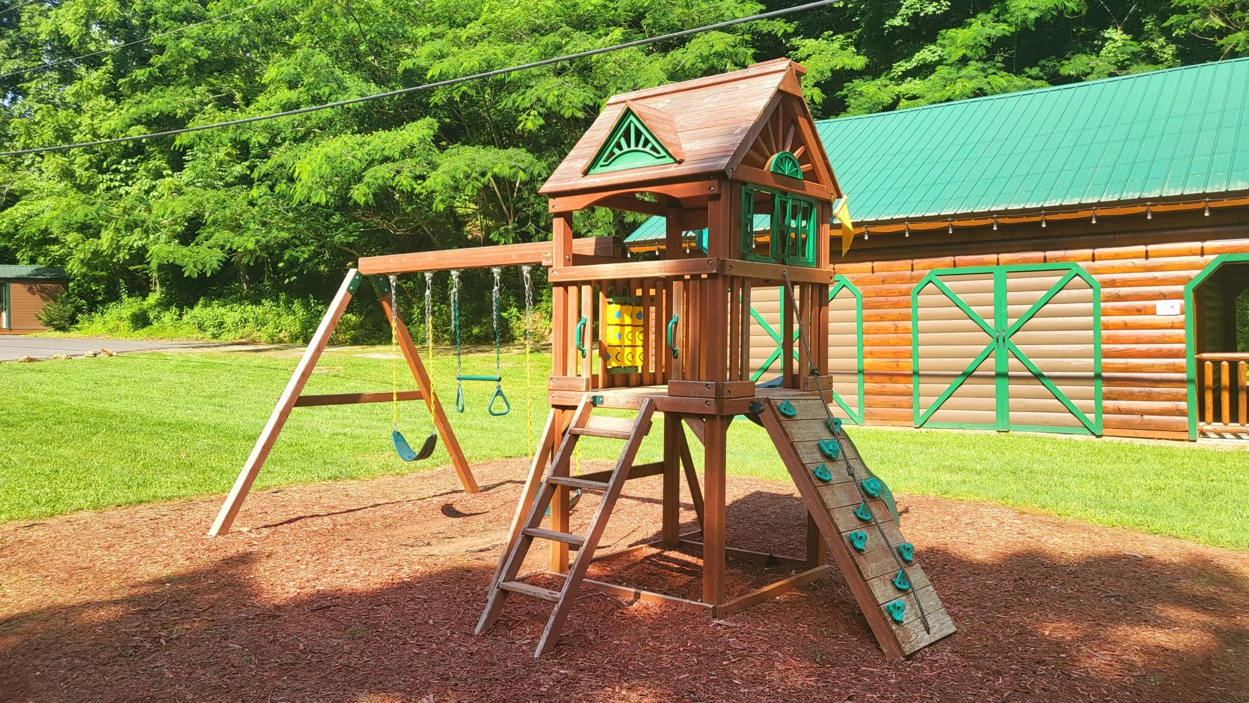 A wooden playground set with swings and a climbing wall on mulch.