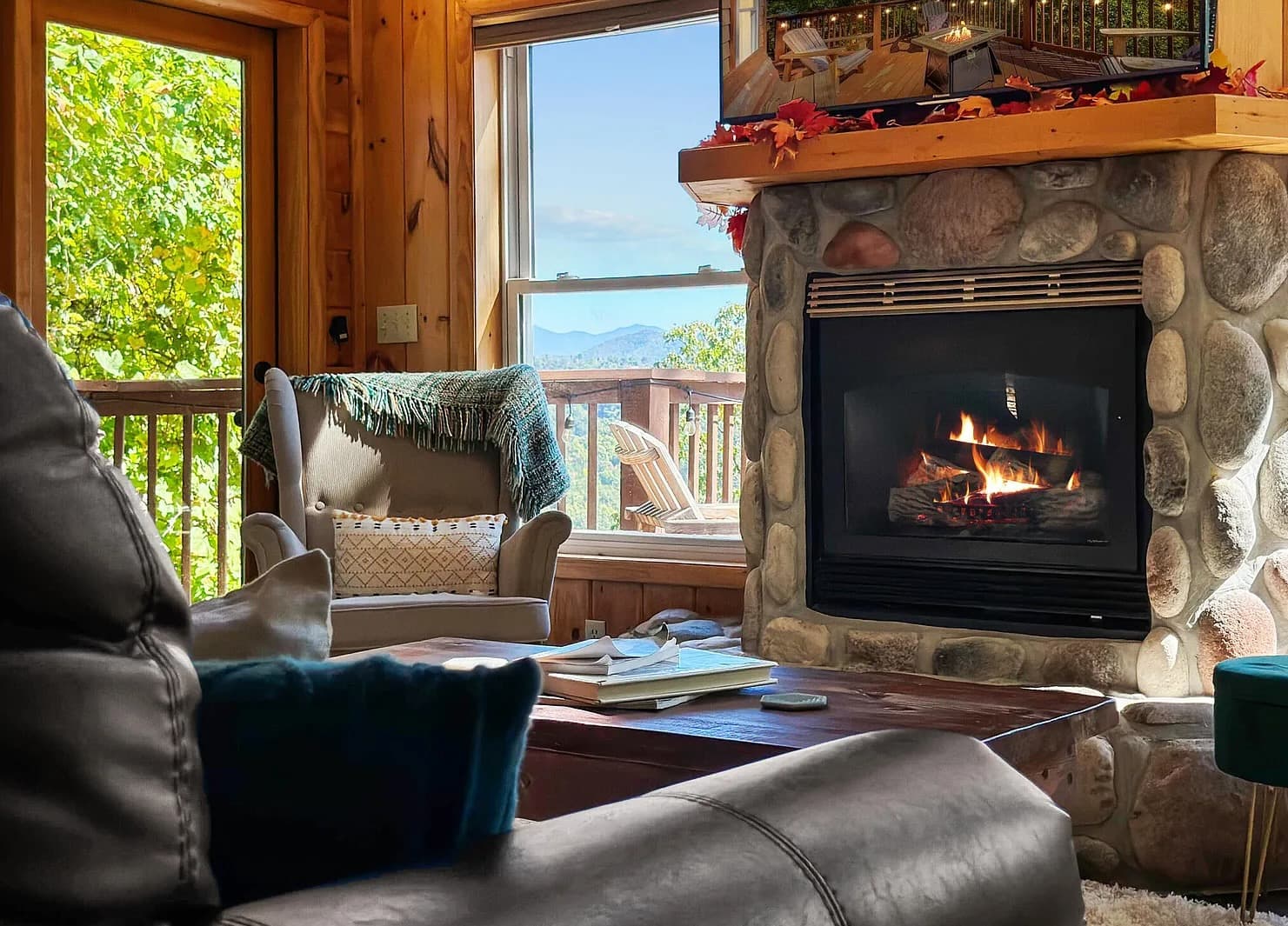 A lit stone fireplace beside a window showing a distant mountain ridge, with a book and remote resting on the coffee table.