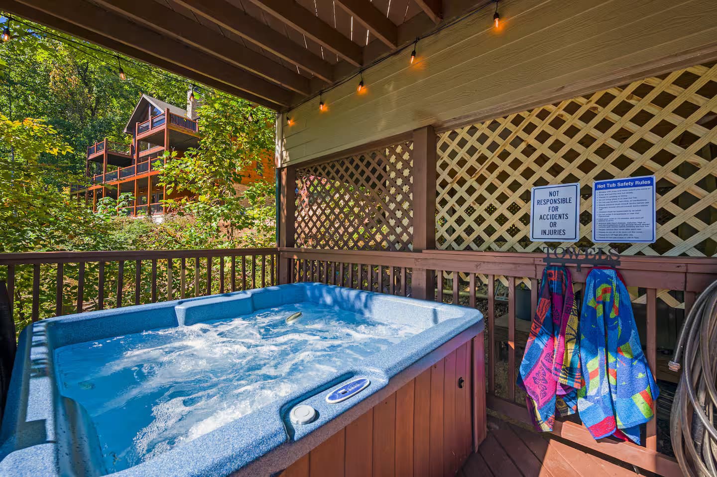 A bubbling hot tub on a covered wooden deck with string lights, towels, and a neighboring cabin visible through the trees.