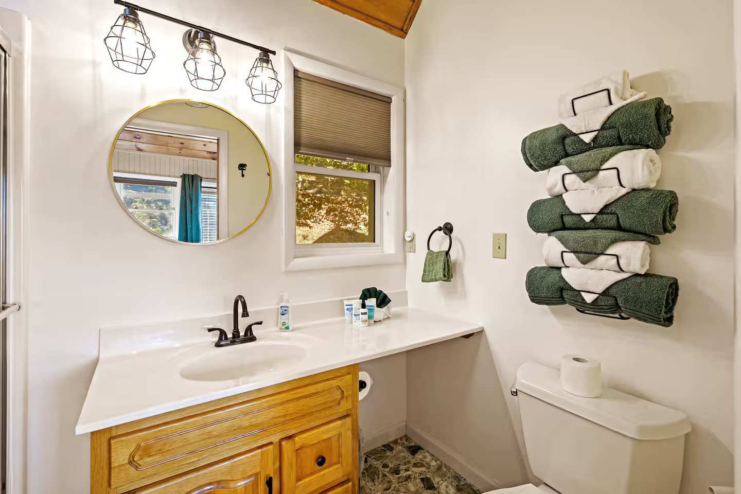 The ensuite bathroom vanity with a round mirror, window, folded towels, and provided toiletries.