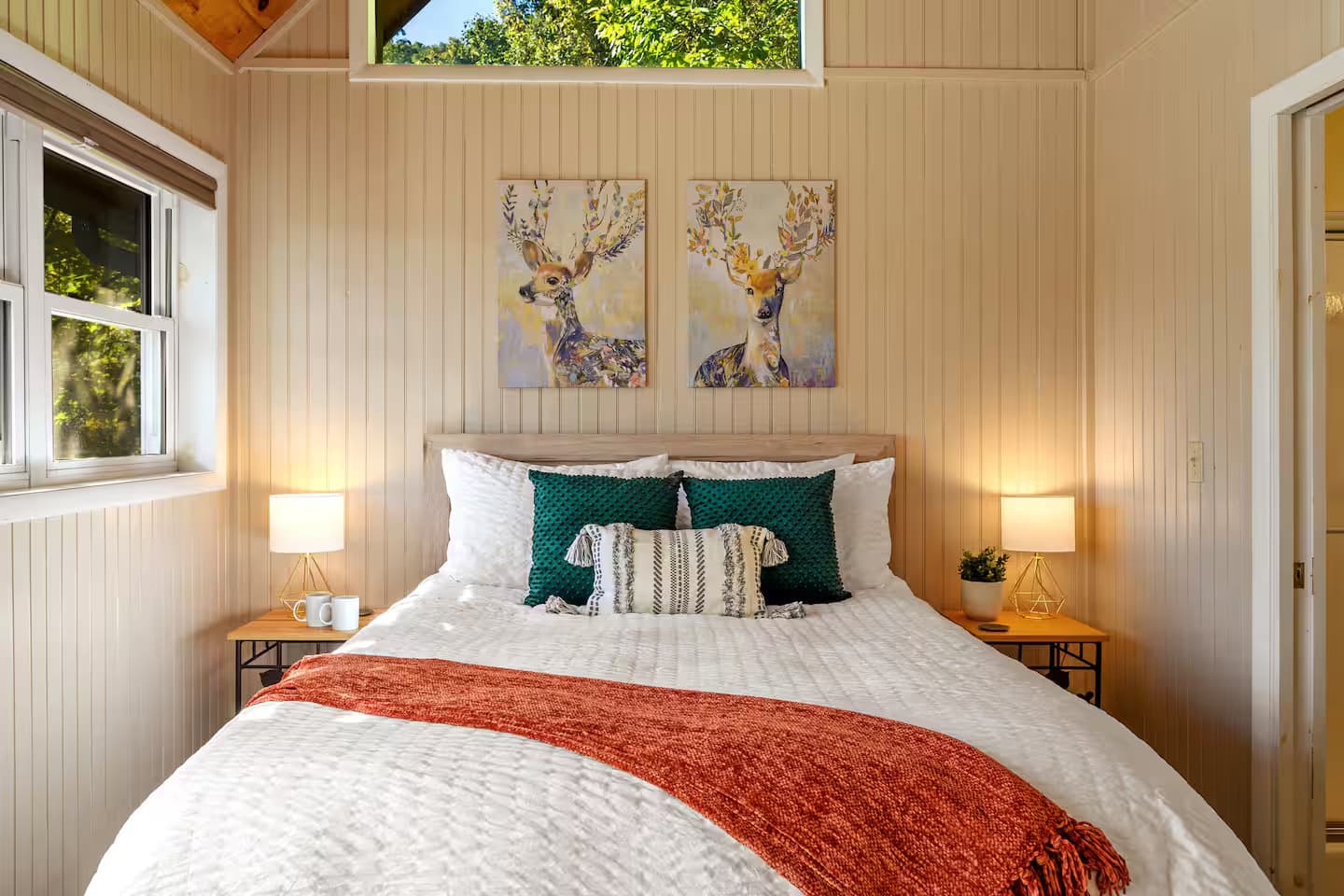 A bedroom with a bed dressed in teal pillows and a rust-colored throw, with matching nightstands and lamps.