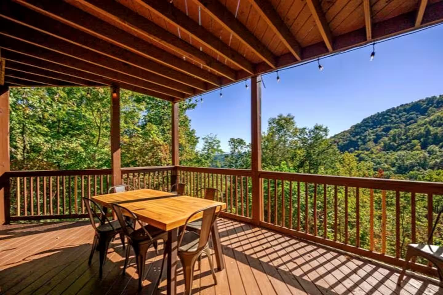 A covered deck dining area with a wood table and metal chairs, overlooking a mountain ridge.