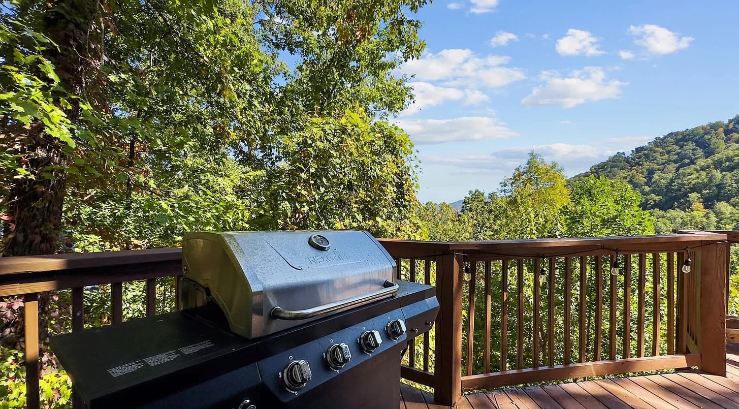 A gas grill on a wraparound deck with string lights overhead and layered green mountain ridges in the background.