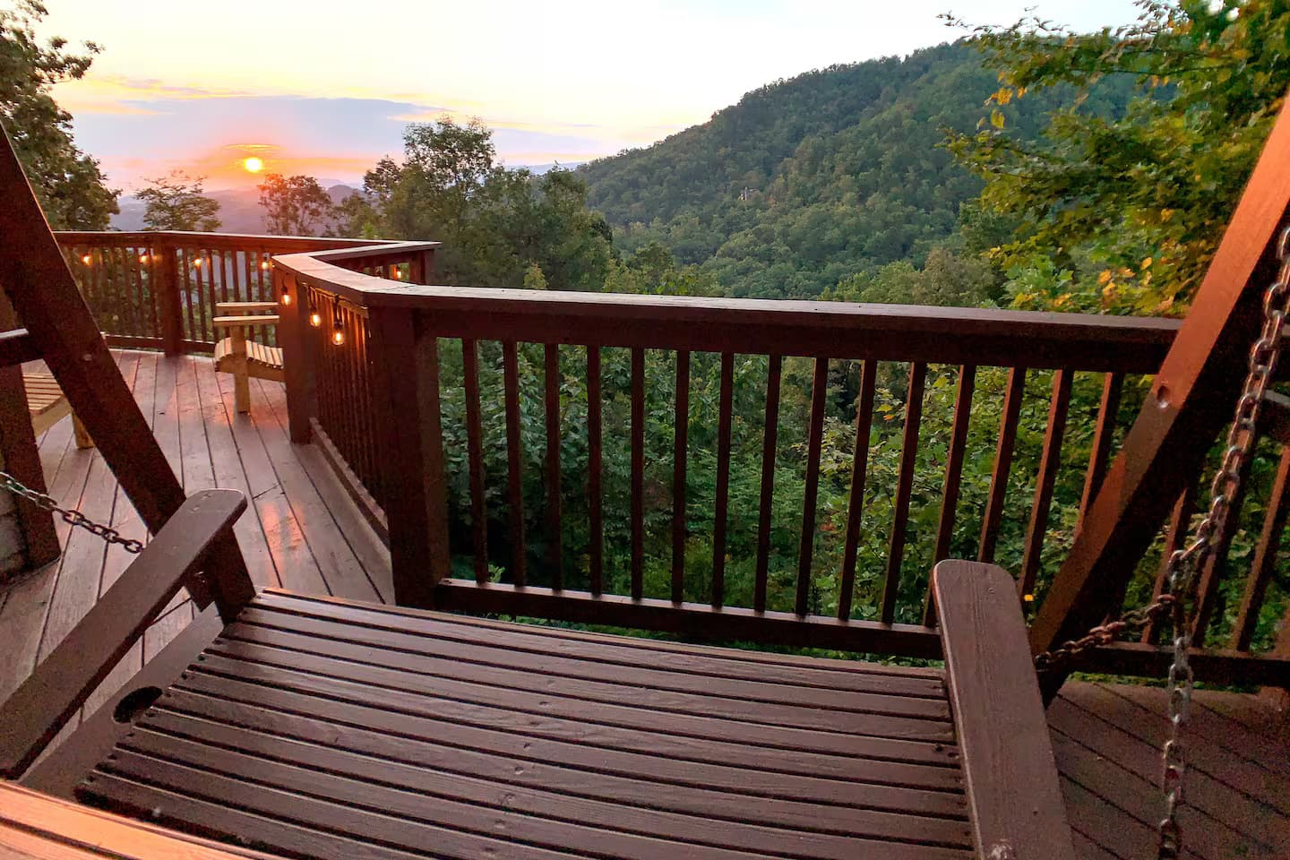 A wooden porch swing on the deck at sunset, with mountain ridges silhouetted against an orange sky.