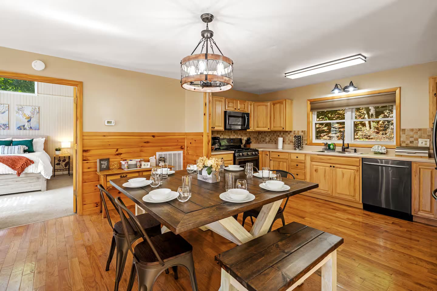 A farmhouse dining table set for six next to a full wood-cabinet kitchen with black appliances.