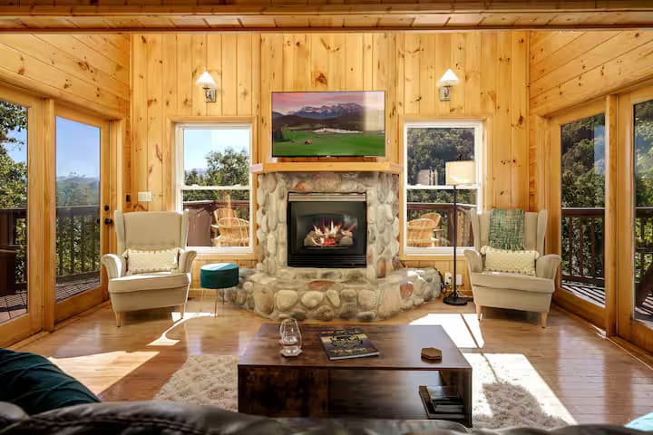 A stone fireplace flanked by two wingback chairs, with mountain views through windows on either side.