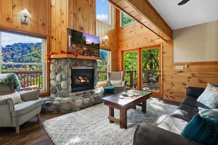 A vaulted living room with a stone fireplace, a wall-mounted TV, and mountain views through windows on two sides.