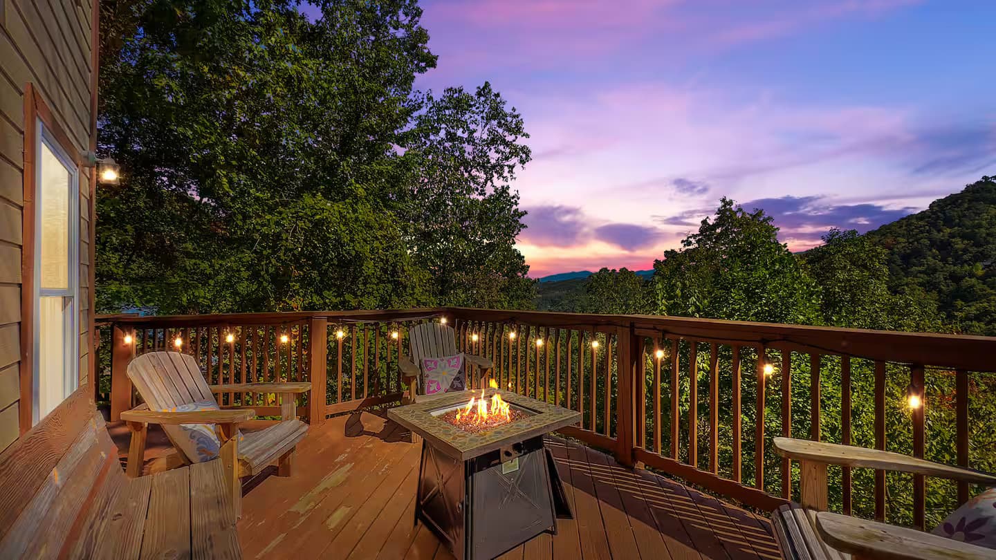 A lit gas fire pit and Adirondack chairs on the cabin deck beneath a pink-and-purple sunset sky.