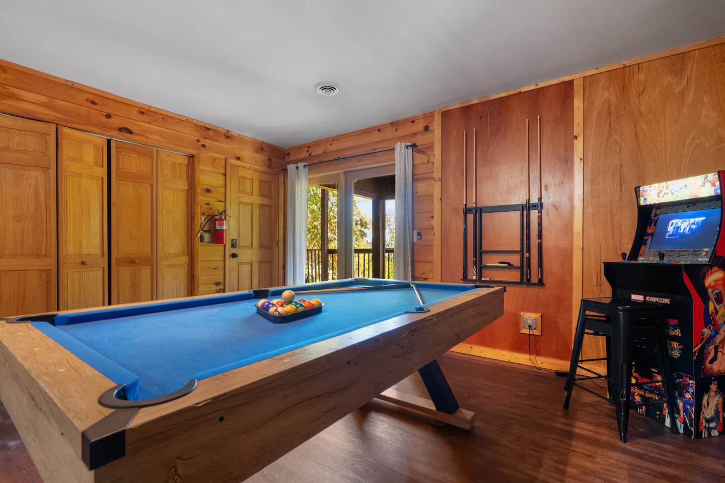 A blue-felt pool table with racked balls beside a standing arcade cabinet and a door opening onto the deck.